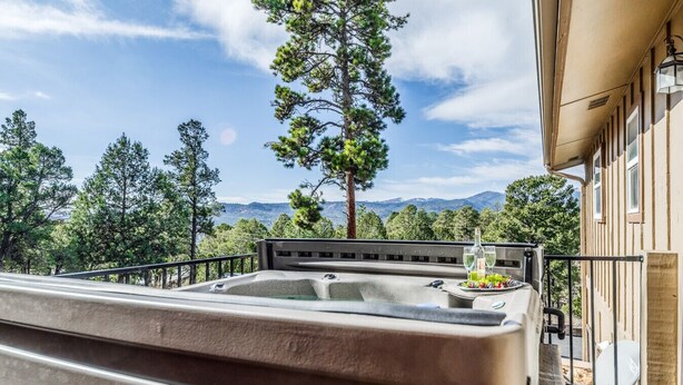 A hot tub on a balcony with a mountain view, a dining table with a bottle and glasses, and a tall pine tree. - Wine & Wilderness: MTN Luxe Chalet/ Hot Tub/ Views (Ruidoso)