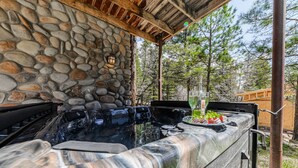 An outdoor kitchen with a stone wall, a grill, and a glass of wine. - Bearfoot Lodge: Hot Tub, Pets, River & Trails (Ruidoso)