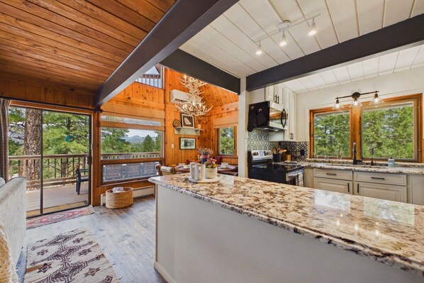 A spacious kitchen with a large island, wooden ceiling, and large windows offering a view of the outdoors.