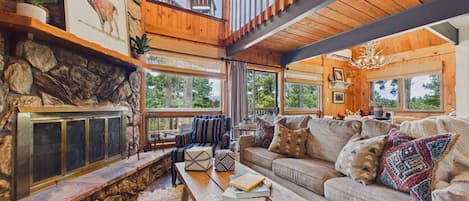 A cozy cabin interior with a stone fireplace, a wooden staircase, a sofa set, and a dining area.