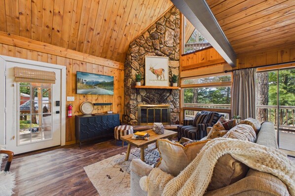 Interior acogedor de una cabaña con chimenea de piedra, paredes de madera y un ventanal con vistas al exterior.