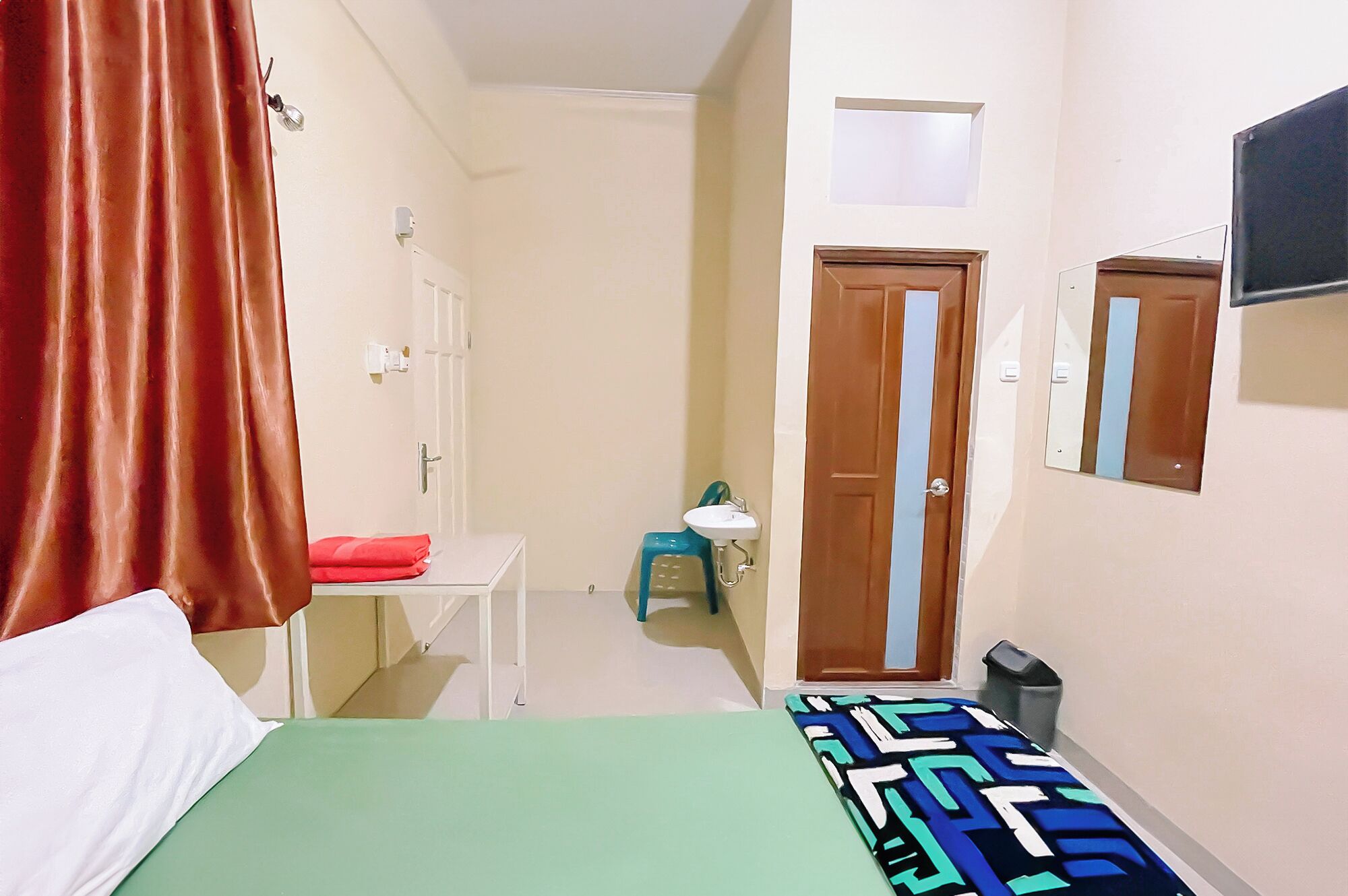 Une petite pièce étroite, comprenant un lit, un bureau, une chaise, un petit lavabo et un miroir.