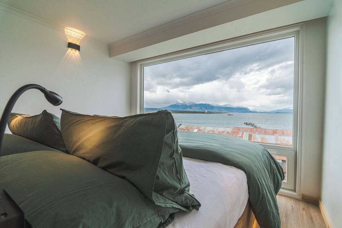 A bedroom with a large window overlooking a body of water and mountains.