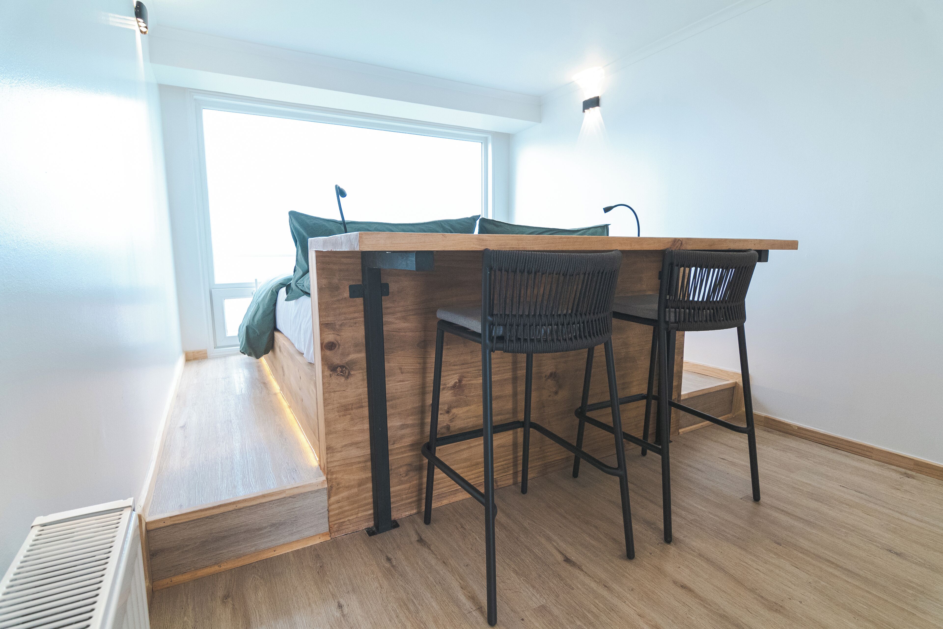 Een kleine kamer met een houten bureau en twee stoelen, een bed met een groene dekbedovertrek, en een raam dat voor natuurlijk licht zorgt.