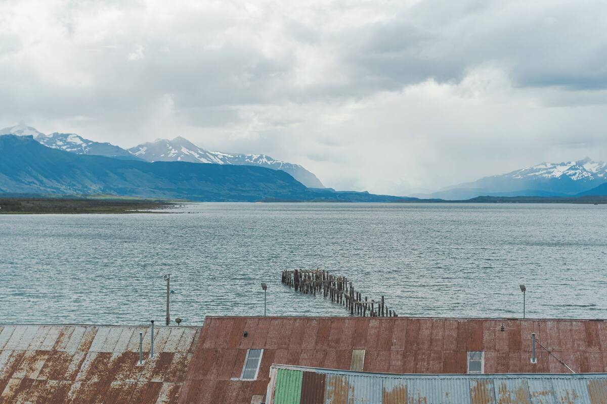 A coastal view with a pier extending into the water, mountains in the background, and a building with a rusted roof.