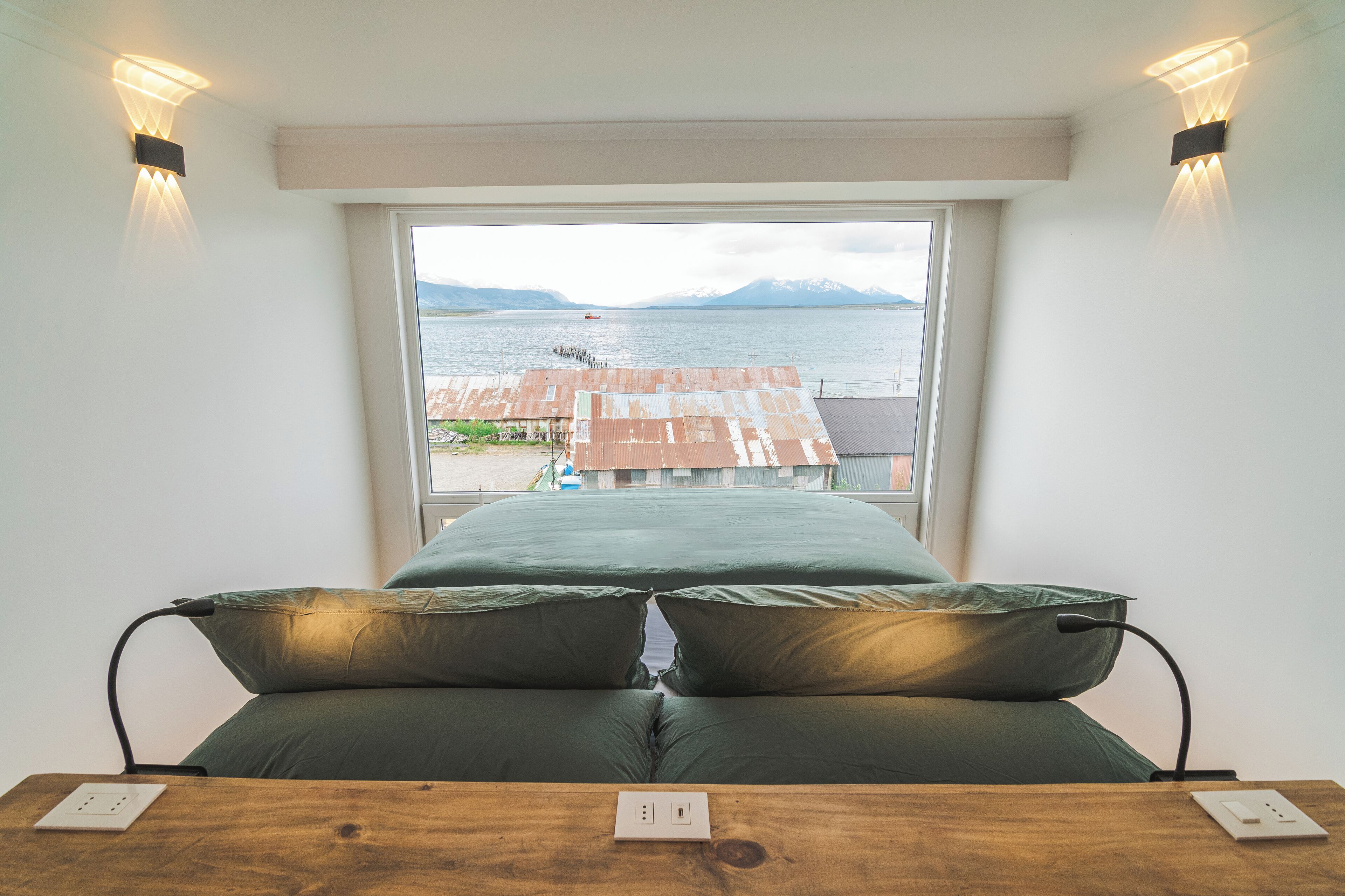 Un dormitorio con un amplio ventanal que da a un cuerpo de agua y montañas, una mesita de noche de madera y una colcha verde.