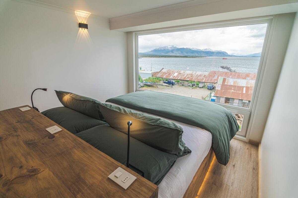 A bedroom with a large window overlooking a body of water and mountains, a wooden bedside table, and a green bedspread.