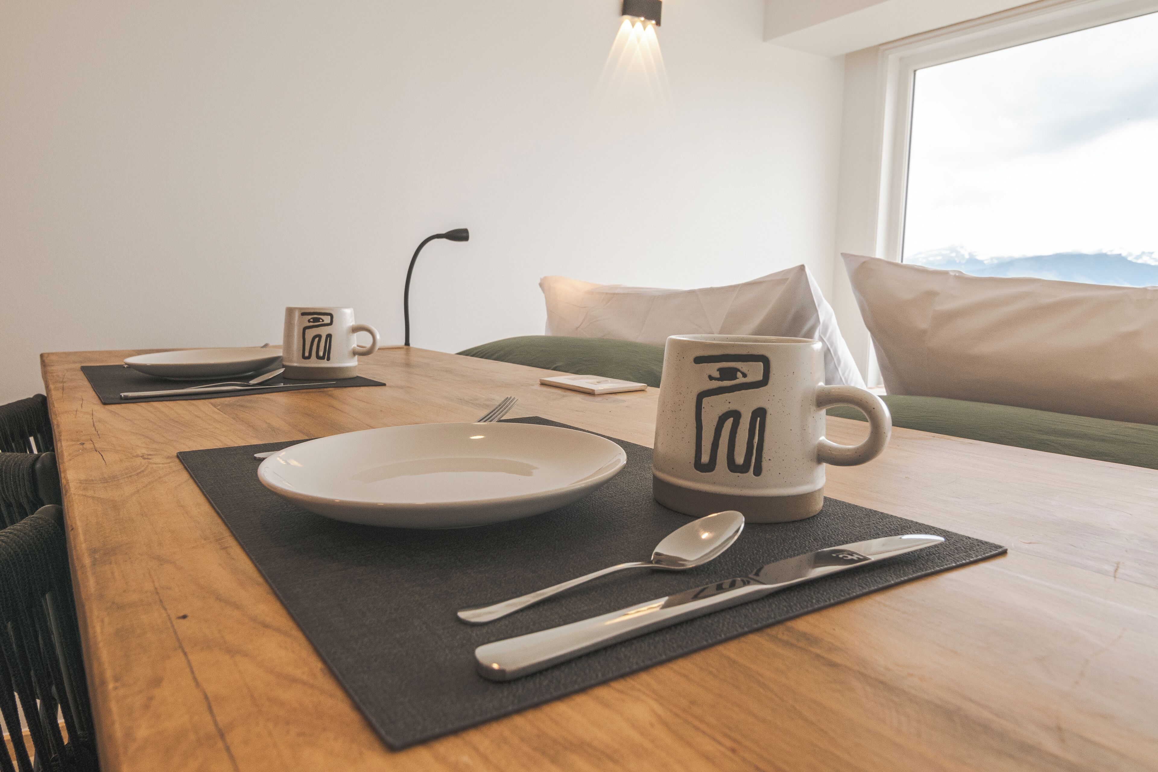 A dining table set with plates, cutlery, and a mug, with a view of mountains through a window.