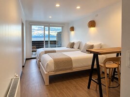A hotel room with two beds, a wooden table, a chair, and a view of mountains through large windows.