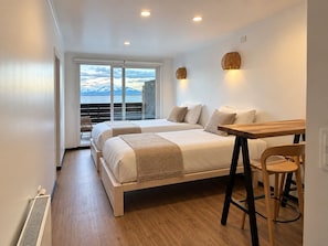 Habitación de hotel con dos camas, una mesa de madera, una silla y vistas a montañas a través de amplias ventanas.
