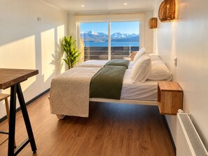 A bedroom with a bed, a wooden table, a potted plant, and a view of mountains through a window. - Loft By Toore Patagonia (Natales)