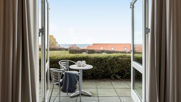 Uitzicht vanaf een balkon met een tafel en stoelen, een zeegezicht en een gebouw met rode daken in de verte.