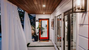 Un corredor cubierto que conduce a una puerta con marco rojo, paredes blancas y techo de madera.