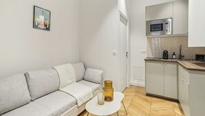 Un appartamento moderno con un divano grigio, un piccolo tavolo bianco, una cucina con forno a microonde e una porta che dà su un'altra stanza.
