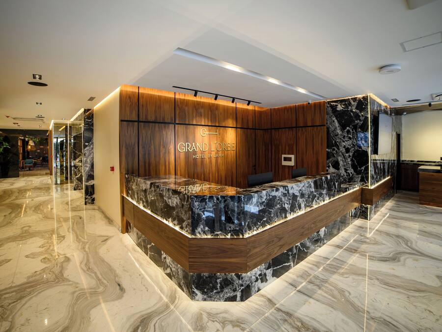 Uma área de recepção de hotel com balcão de mármore, painéis de madeira e uma placa com a inscrição 'GRAND LORBE HOTEL BY JAM'.