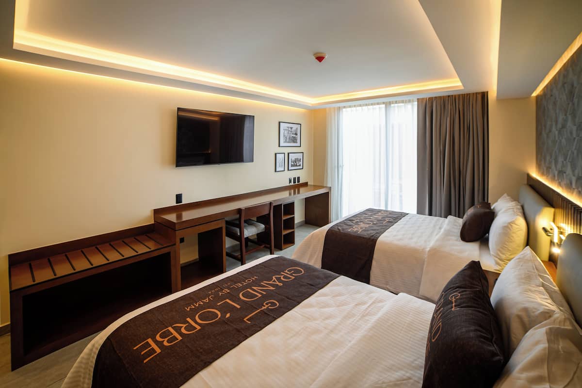 A hotel room with two beds, a desk, a flat-screen TV mounted on the wall, and a window with curtains.