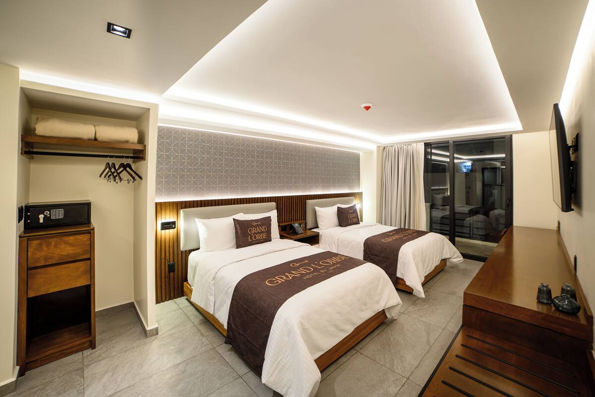 A hotel room with two beds, a desk, a wardrobe, and a bathroom visible through a glass door.