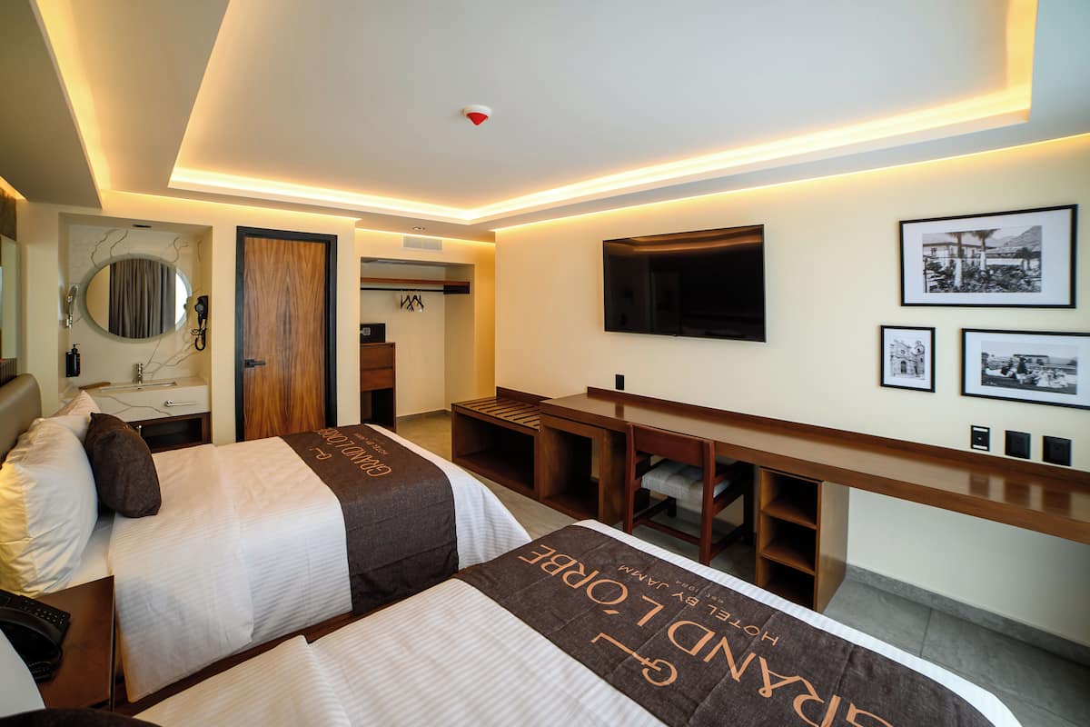 A hotel room with two beds, a desk, a TV, and framed pictures on the wall.