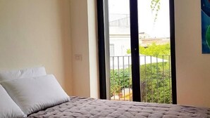A bedroom with a bed, white pillows, and a view of a balcony with greenery and a building in the background. - The Sicilian (Acireale)