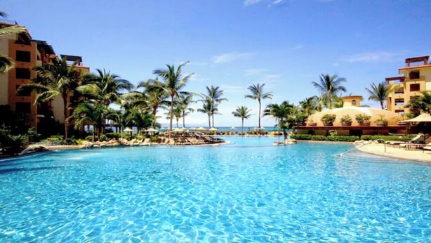 A large, clear swimming pool with palm trees and resort buildings in the background.