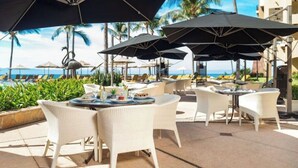 An outdoor seating area with white wicker chairs and tables, umbrellas, and a view of the ocean.