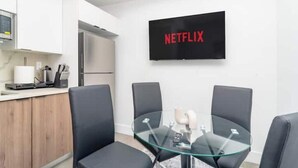 Een moderne keuken met witte kastjes, een magnetron en een koffiezetapparaat. Er staat een glazen tafel met vier zwarte stoelen en een televisie aan de muur met daarop 