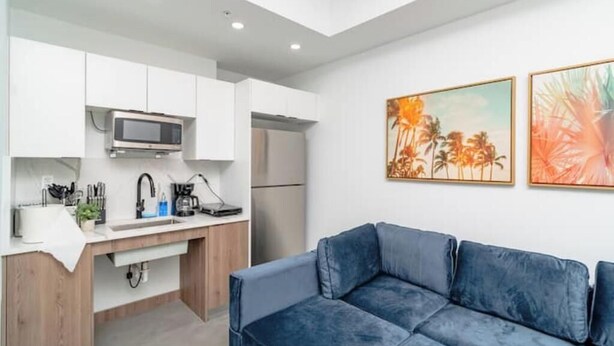 A modern kitchen with white cabinets, a microwave, and a refrigerator. There is a blue sofa in the living area and two framed artworks on the wall. - King Bed brand new safe Oasis minutes to Brickell (Miami)
