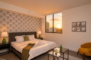 Hotelkamer met een bed, twee bedlampen, een glazen salontafel, een stoel en een raam met uitzicht op een zonsondergang.