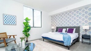 A modern bedroom with a bed, bedside tables, a chair, a glass coffee table, and a large window.