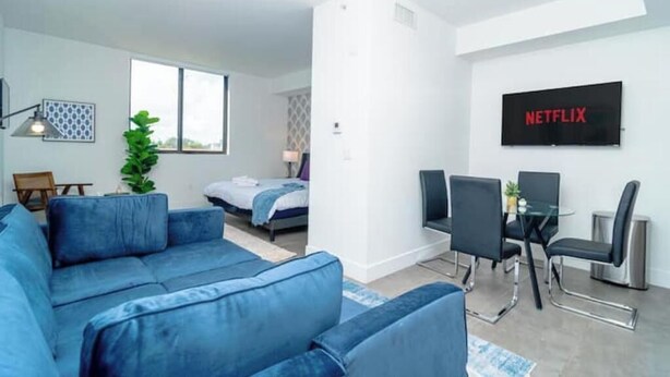 A modern living room with a blue sofa, a dining area with a glass table and chairs, a bed, and a large window.