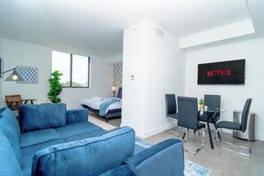 Een moderne woonkamer met een blauwe bank, een eethoek met een glazen tafel en stoelen, een bed en een groot raam.