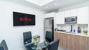 A modern kitchen with white cabinets, a microwave, and a refrigerator. There is a glass table with chairs and a small plant. A TV mounted on the wall displays the Netflix logo.