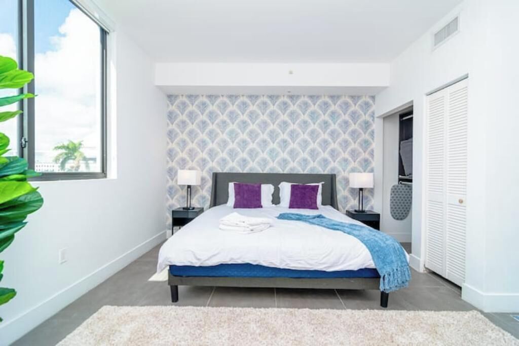 A modern bedroom with a bed, two bedside tables, a nightstand, a large window, and a view of the outdoors.