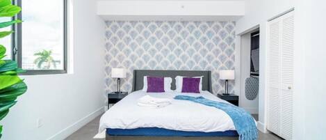 A modern bedroom with a bed, two bedside tables, a nightstand, a large window, and a view of the outdoors.