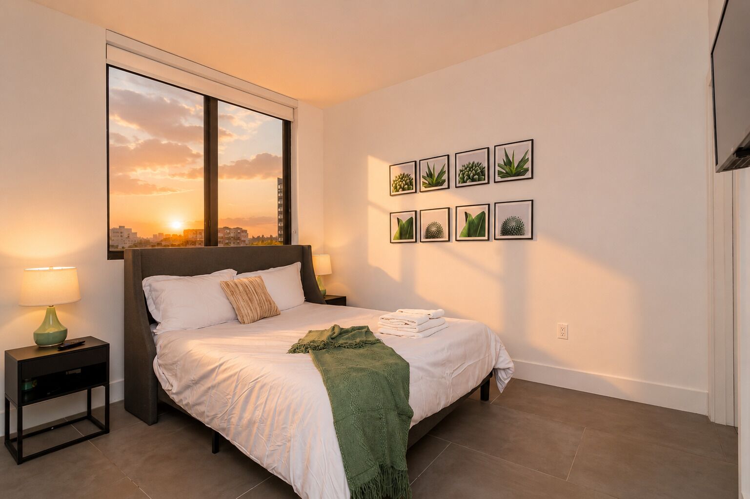 A bedroom with a bed, a nightstand, a window with a sunset view, and wall art.