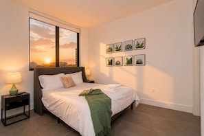 Une chambre à coucher avec un lit, une table de chevet, une fenêtre donnant sur un coucher de soleil et des œuvres d’art murales.