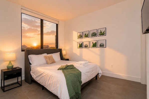 A bedroom with a bed, a nightstand, a window with a sunset view, and wall art.
