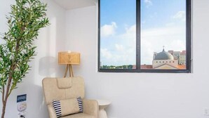Ein Zimmer mit großem Fenster, einem Sessel, einem kleinen Tisch und einer Lampe.