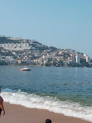 A coastal cityscape with buildings on a hillside, a body of water with buoys, and a beach with waves.