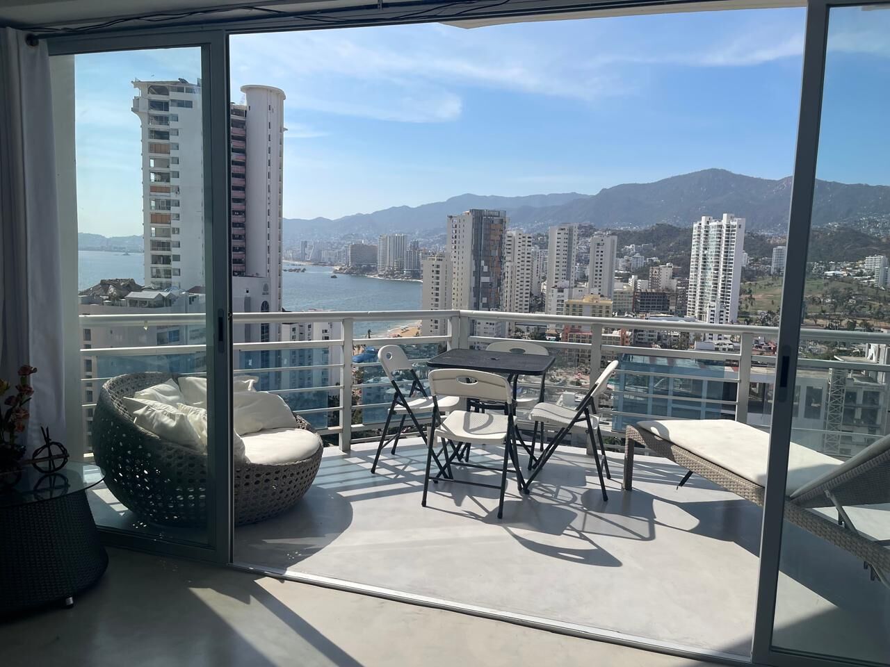 En balkon med et bord og stole, en fletstol og udsigt til by og bjerge.
