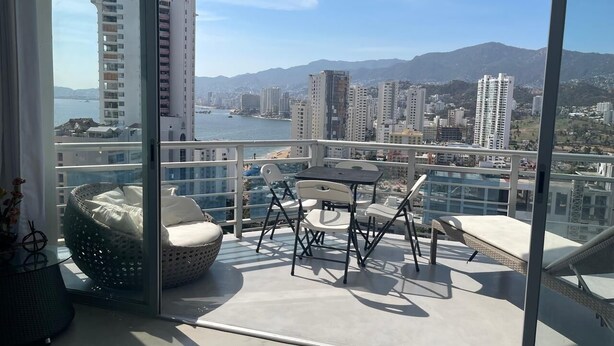 A balcony with a table and chairs, a wicker chair, and a view of a cityscape and mountains. - Apartment with direct access to the beach, fully equipped and renovated. (Acapulco de Juárez)