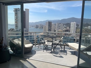 A balcony with a table and chairs, a wicker chair, and a view of a cityscape and mountains.