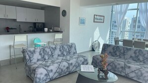 A modern living room with marble-patterned sofas, a glass coffee table, and a kitchen area with white cabinets and a clock. - Apartment with direct access to the beach, fully equipped and renovated. (Acapulco de Juárez)