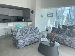 A modern living room with marble-patterned sofas, a glass coffee table, and a kitchen area with white cabinets and a clock. - Apartment with direct access to the beach, fully equipped and renovated. (Acapulco de Juárez)