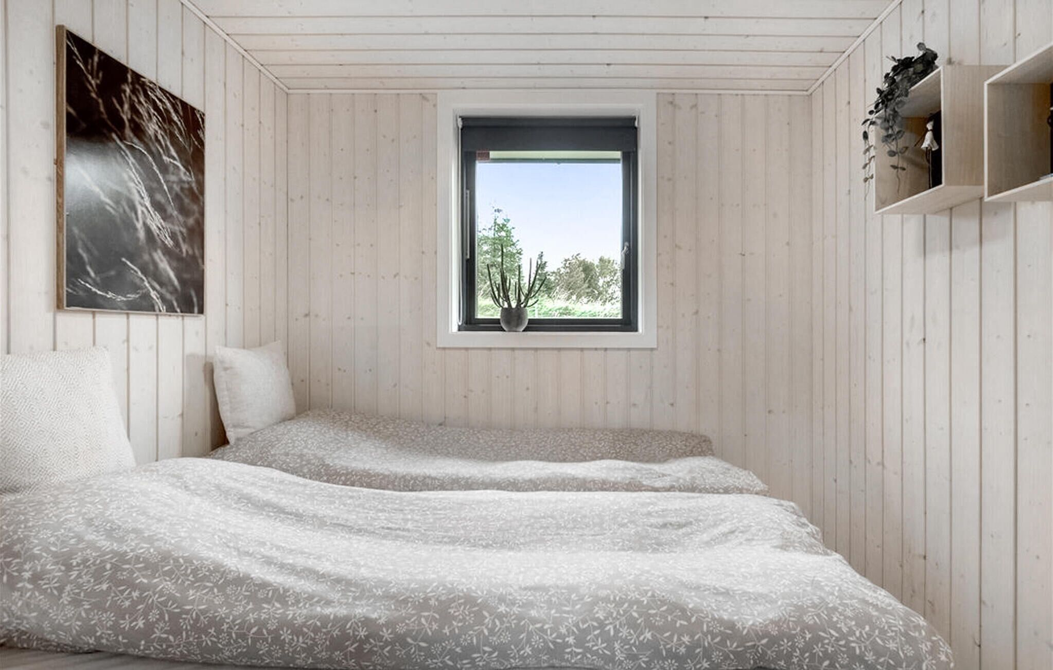 A bedroom with two beds, a window with a view of greenery, a wall-mounted shelf with decorative items, and a framed picture on the wall.