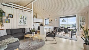 Un salon moderne comprenant un canapé, une table basse et des fauteuils, un coin cuisine avec une table à manger, et de grandes fenêtres offrant une vue sur l’extérieur.