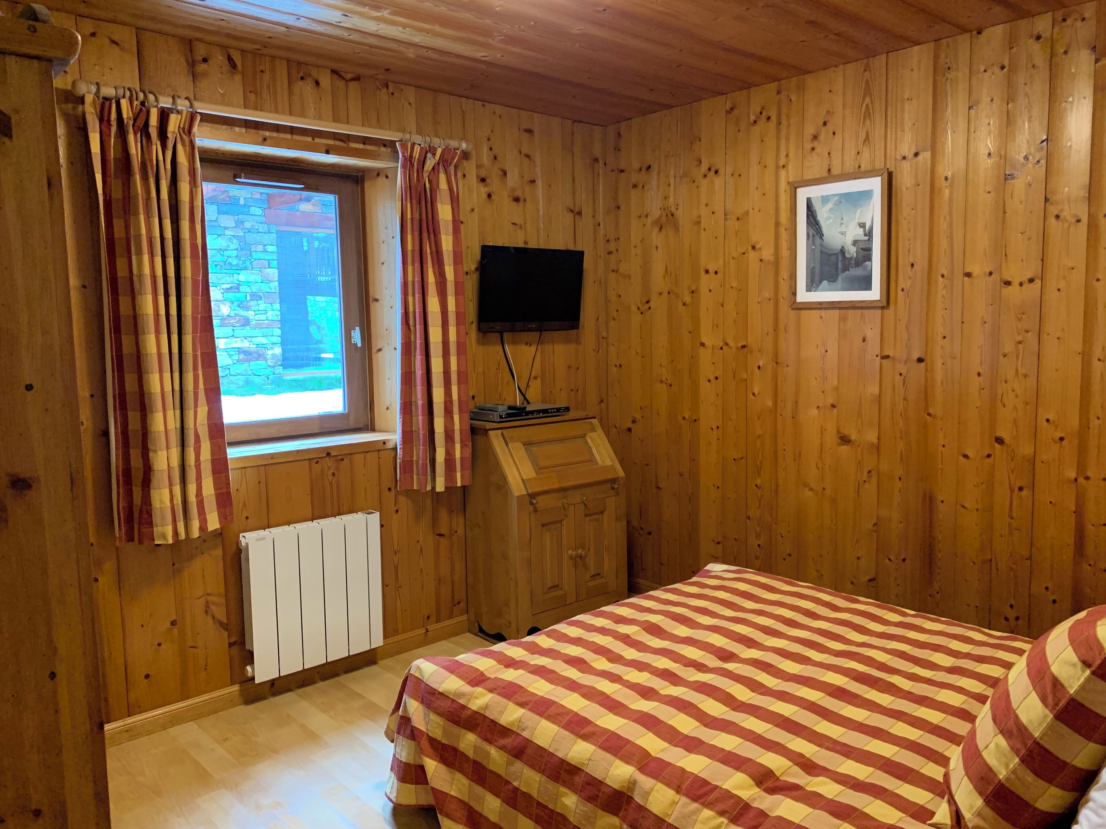 A wooden-paneled room with a bed, a window with curtains, a radiator, a television, and a framed picture on the wall.