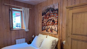Une pièce aux murs lambrissés, comprenant un lit, une fenêtre avec des rideaux, un radiateur, une table de chevet avec une lampe et une tapisserie représentant une scène de village.