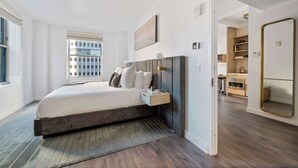 A modern bedroom with a large bed, a bedside table, a mirror, and a view of the city through the window. - 17John: Luxury One Bedroom Apartment (New York)