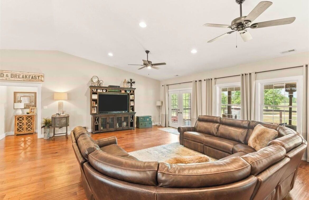 A spacious living room with a leather sectional sofa, a flat-screen TV, and ceiling fans.